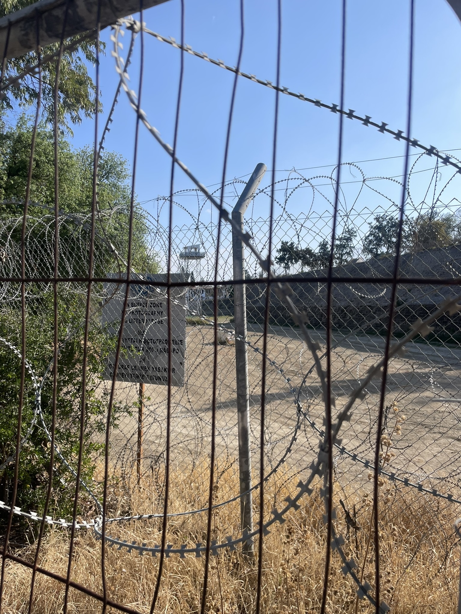 Multiple layers of security fencing and barbed wire on the approach to the Ledra Palace checkpoint, with a watch post and tennis court behind.